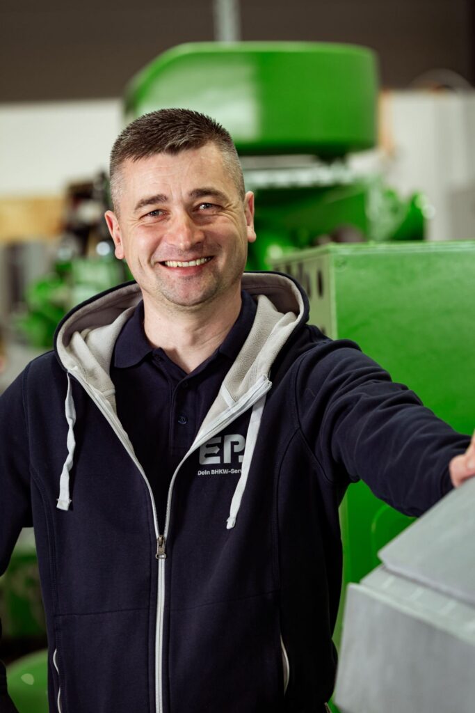 Mann mit kurzen dunklen Haaren, lacht und steht an einem grünen Jenbacher Motor, er trägt einen EPS Hoodie